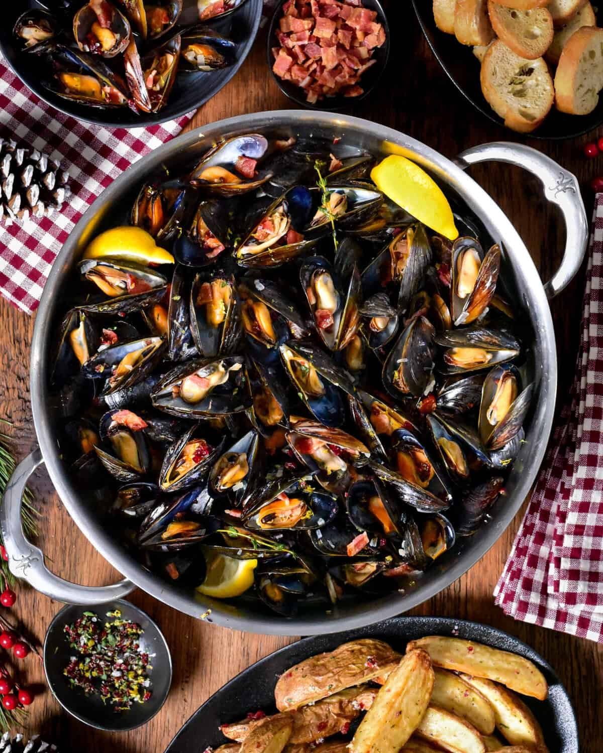 A pot of PEI Mussels in Cider Broth with Bacon and Cranberry Salt. they are served bread, lemon wedges and salt and malt vinegar potato wedges.