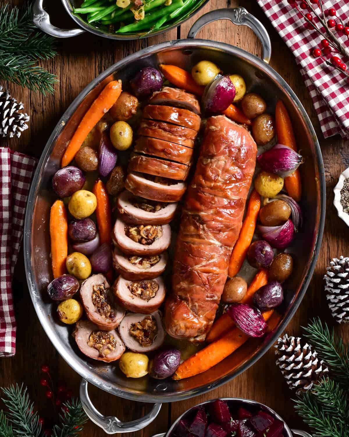 Two Prosciutto Wrapped Pork Tenderloin Stuffed with Figs & Brie (one is sliced) and served in an oval platter with roasted vegetables.