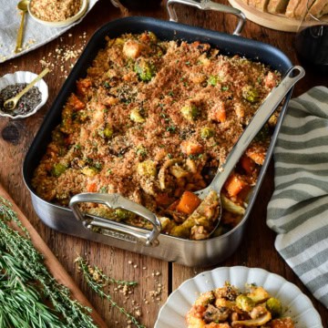 A winter vegetable cassoulet in a roaster with a serving spoon. A portion is served on a plate and accompanied by extra breadcrumb toppings and sourdough bread.