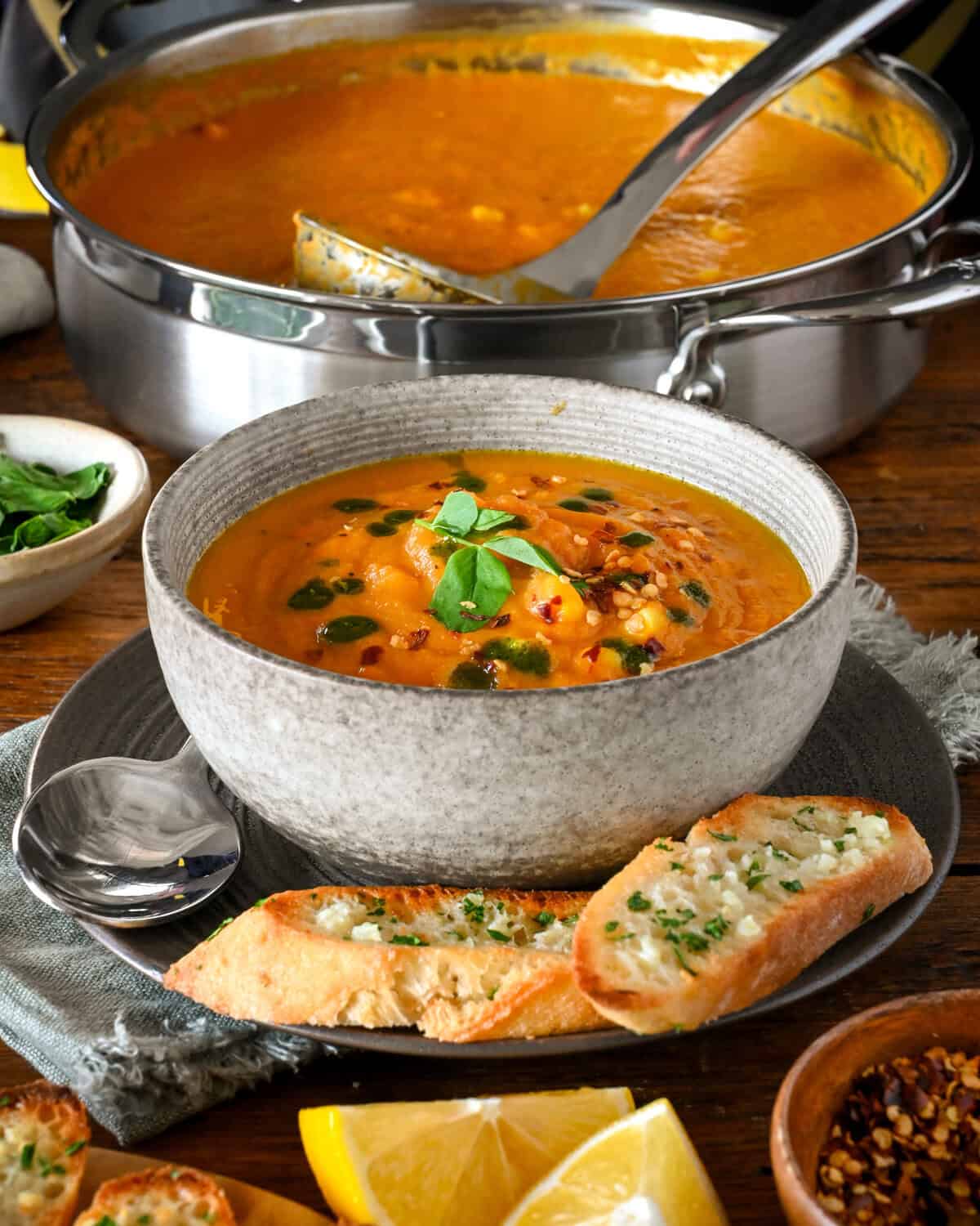 A bowl of roasted carrot chowder with toppings, with a pot in the background. The soup is served with garlic bread.