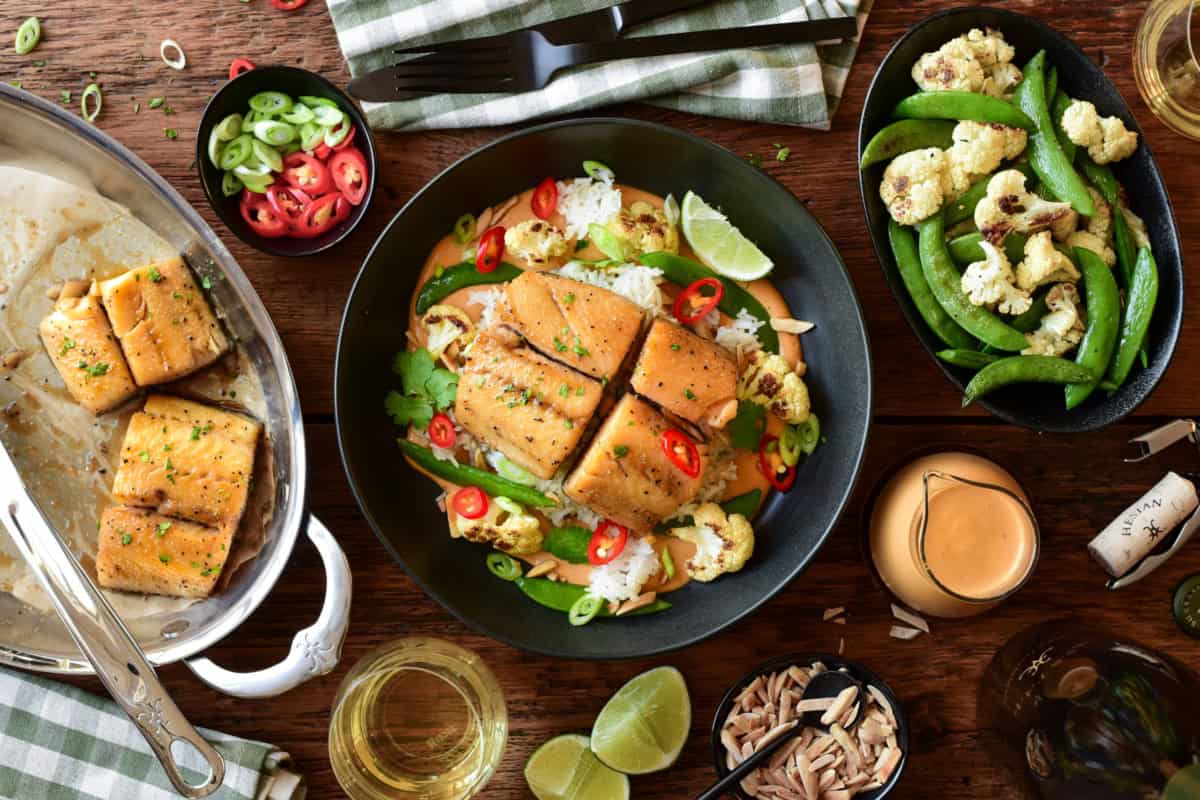 Baked black cod served in a bowl with Thai peanut curry sauce, crispy rice, roasted cauliflower, snap peas, red chilli peppers and a lemon wedge.