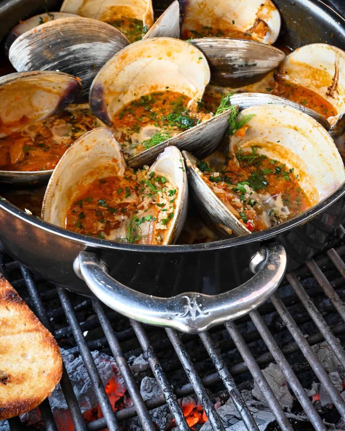 A pot of cherrystone clams in a Calabrian chili sauce over a BBQ with some grilled toast.