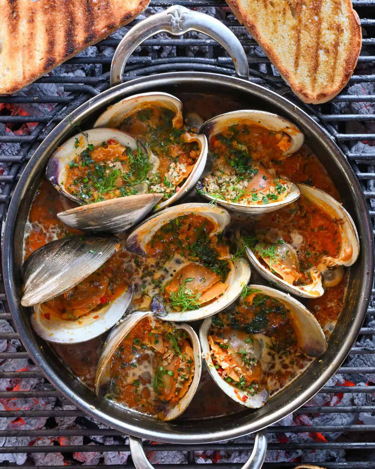 A pot of grilled clams casino in a Calabrian chili sauce over a BBQ with some grilled toast.