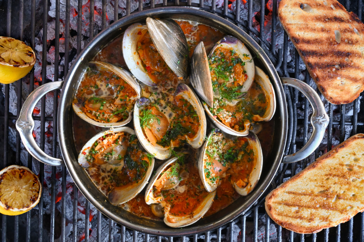 A pot of grilled clams casino in a Calabrian chili sauce over a BBQ with some grilled toast.