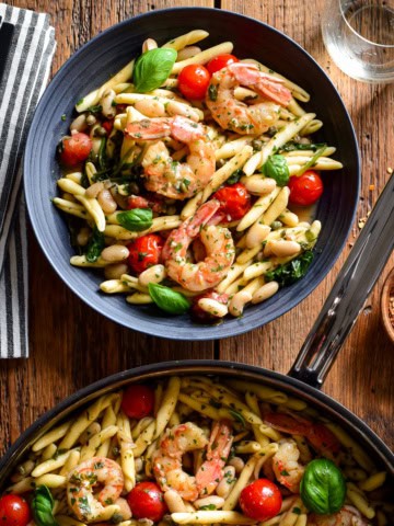 A pan and a bowl with Lemon Caper Shrimp Piccata Pasta with White Beans. This pasta is served with bread, chili flakes white wine an extra fresh basil and tomatoes.