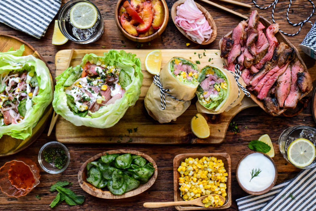 The image features a variety of fresh wraps filled with vegetables and meats, accompanied by grilled lamb slices, a bowl of corn salad, and fresh herbs on a rustic wooden table. Lemon wedges and small bowls of sauces add flavour, creating an inviting and colourful meal presentation ideal for a healthy dining setting at Zimmy's Nook.