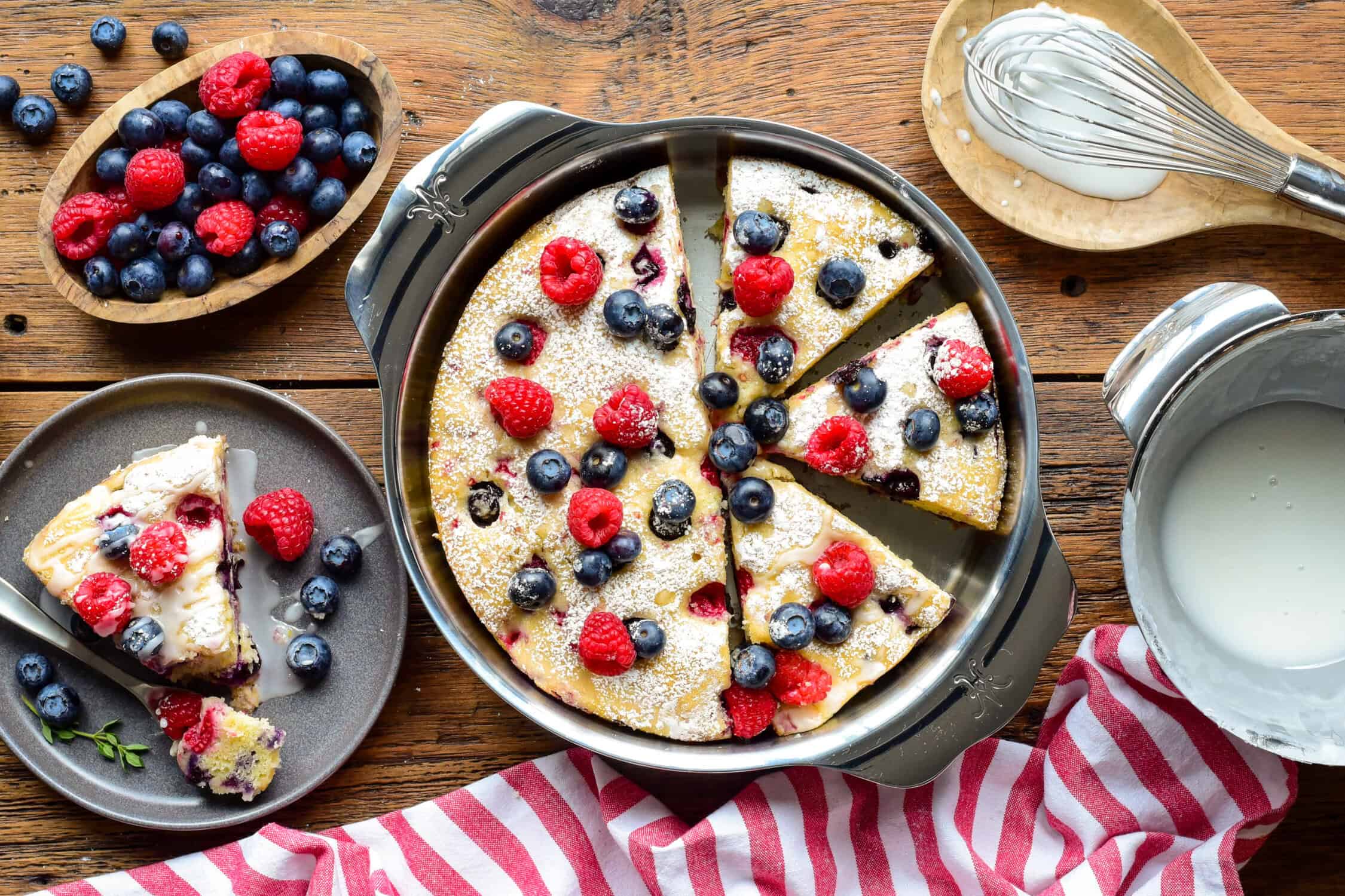 A Lemon Blueberry and Raspberry Cake with blueberries and lemon drizzle on a round serving plate on a rustic wooden table with a red and white striped tea towel. A slice is served with extra berries and glaze, with extra berries on the side.