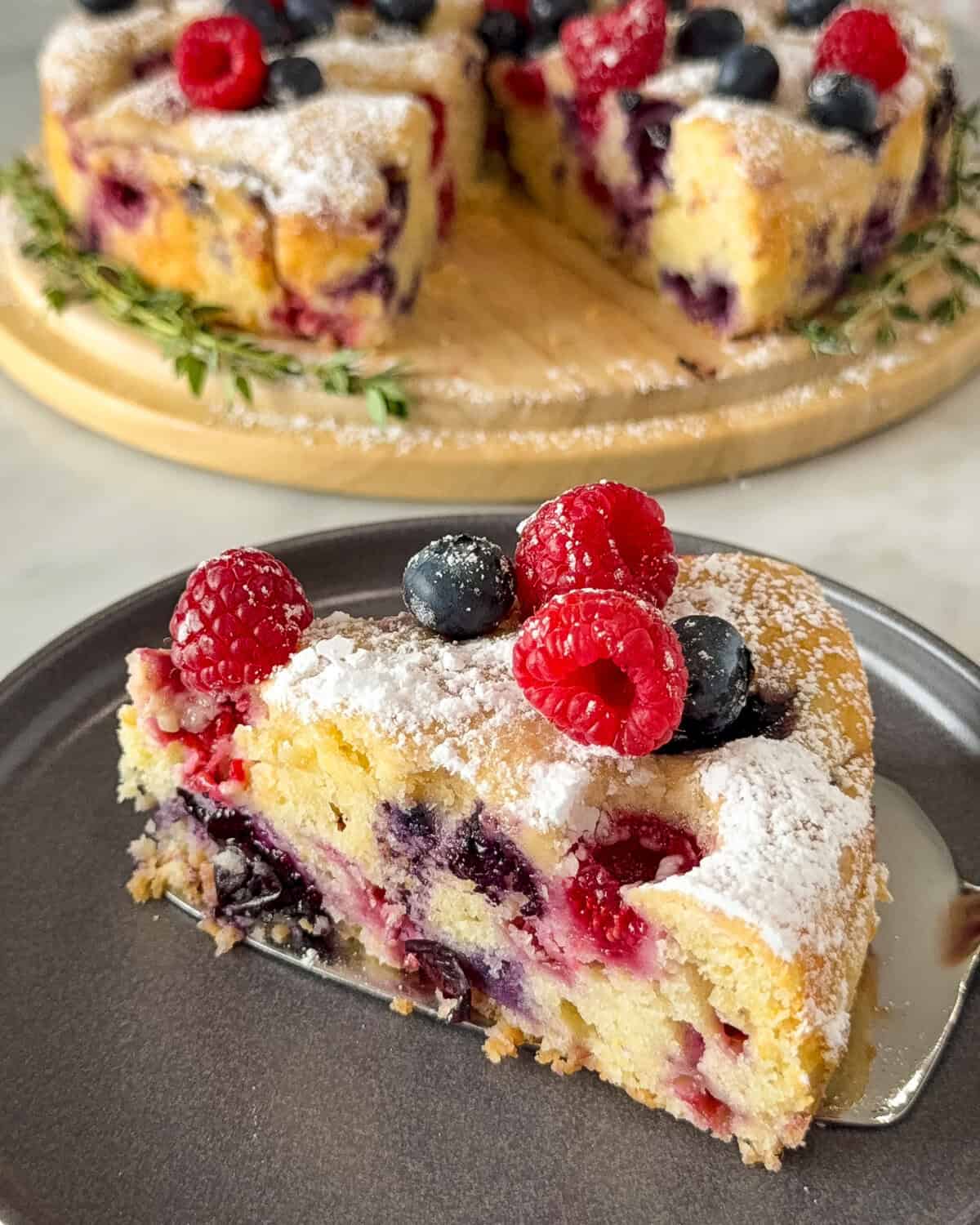 A slice of lemon, blueberry and raspberry cake topped with icing sugar with the cake in the background.