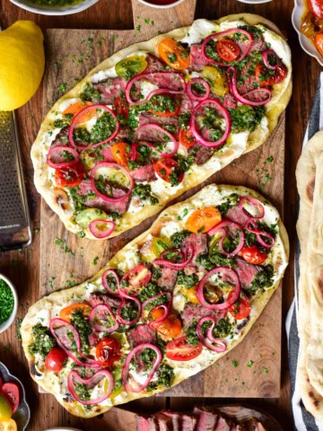 Two chimichurri steak flatbreads with whipped feta, marinated onions and blistered tomatoes on a wooden board. Extra ingredients are placed around this board.
