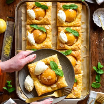 Two hands holding a plate with a hot honey peach puff pastry with a dollop of mascarpone, mint leaves and slivered almonds. It's being held above a sheet pan with two rows of the peach tarts below.