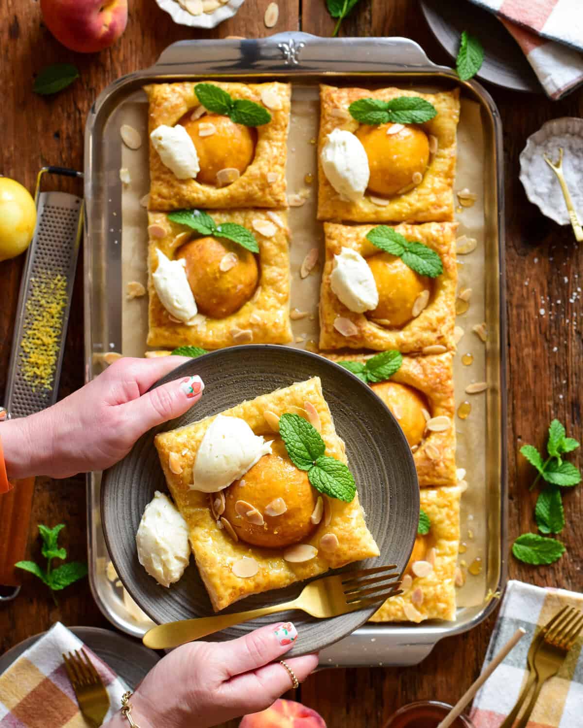 Two hands holding a plate with a hot honey peach puff pastry with a dollop of mascarpone, mint leaves and slivered almonds. It's being held above a sheet pan with two rows of the peach tarts below.