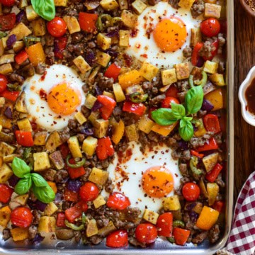 A stainless steel griddle with an Italian sausage and pepper breakfast hash with three fried eggs.