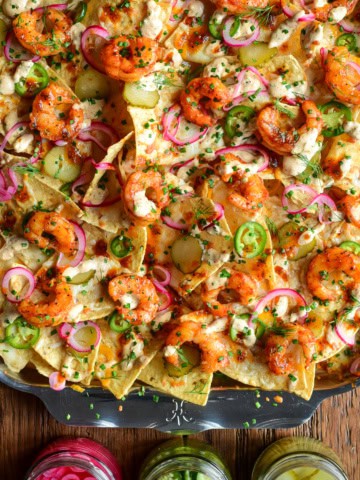 A close-up of a tray with Nashville Hot Honey Shrimp Nachos with Pickle Crema. Layered with pickled onions, jalapeños, dill pickle slices and herbs. Served with extra pickles on the side.