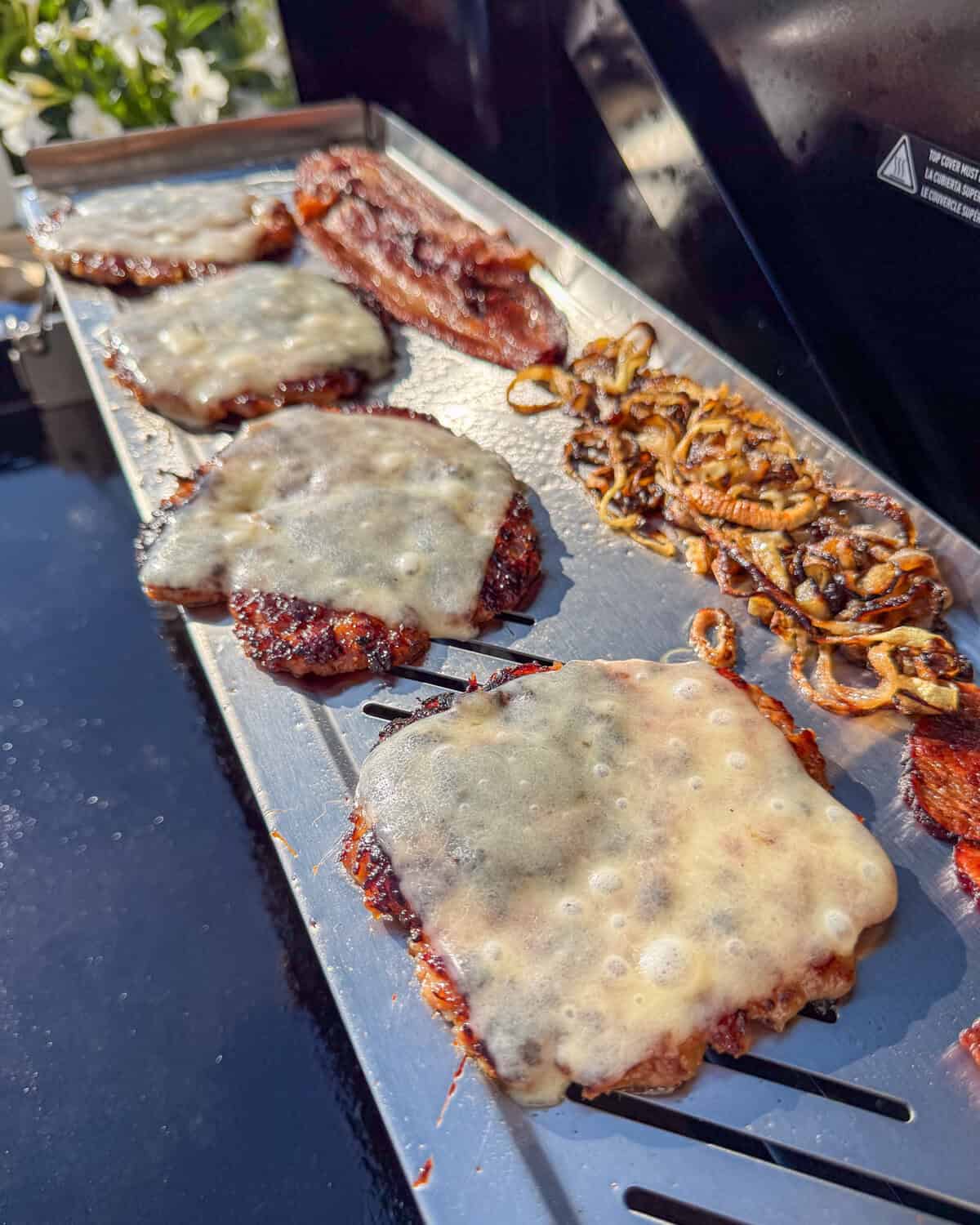 Four cheesy smash burgers on the upper rack of a grill with bacon and fried onions.