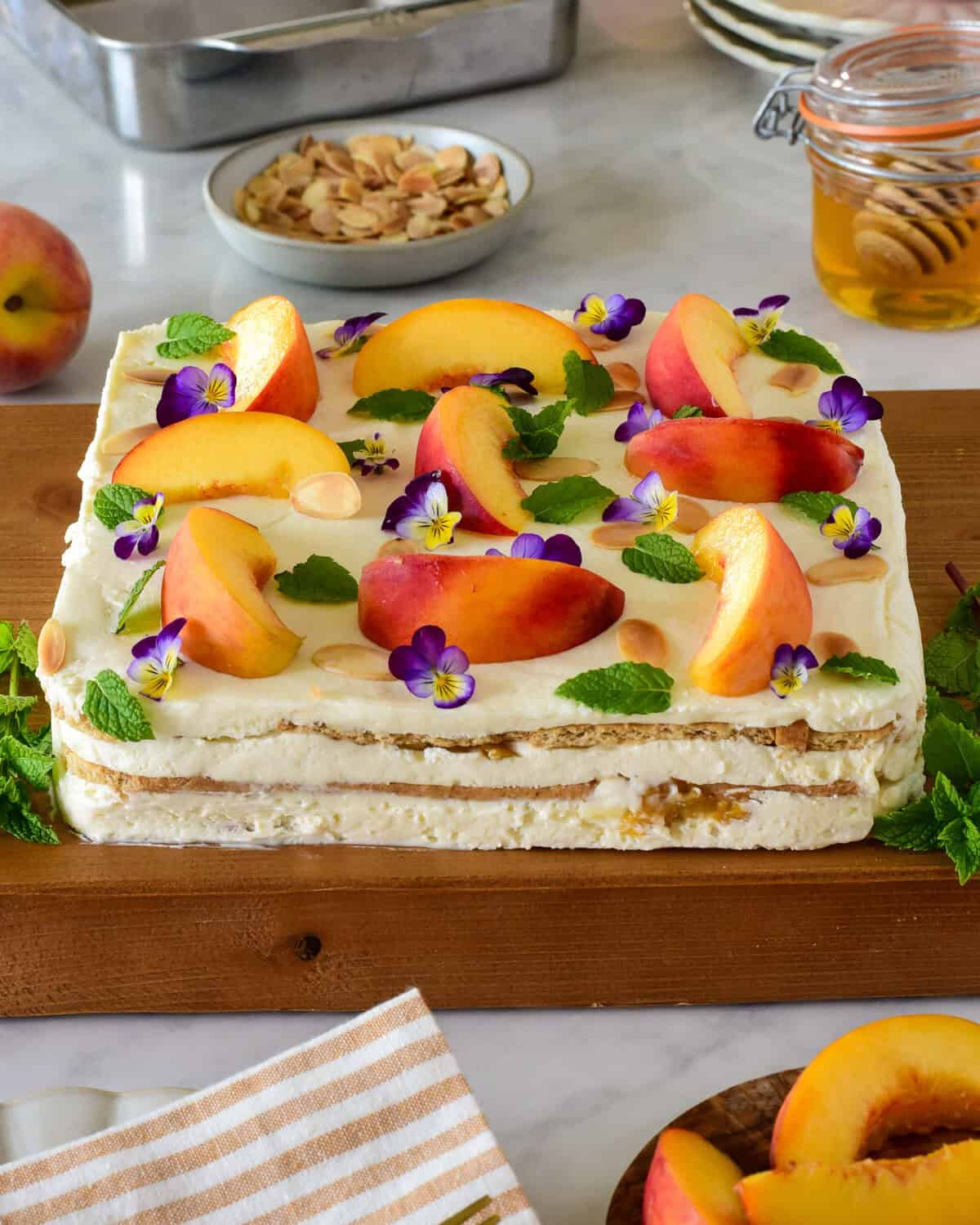 A caramelized peach icebox cake with layers of graham crackers and mascarpone whipped cream. The cake is decorated with fresh peaches, mint and viola edible flowers.