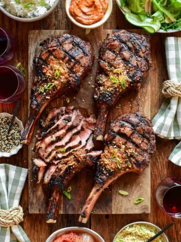 Four veal rib chops with distinct grill marks, one of which has been thinly sliced. They are placed on a wooden cutting board and served with extra gochujang butter, a Korean salad, kimchi and toasted sesame seeds. There are checkered napkins and a few red wine glasses.