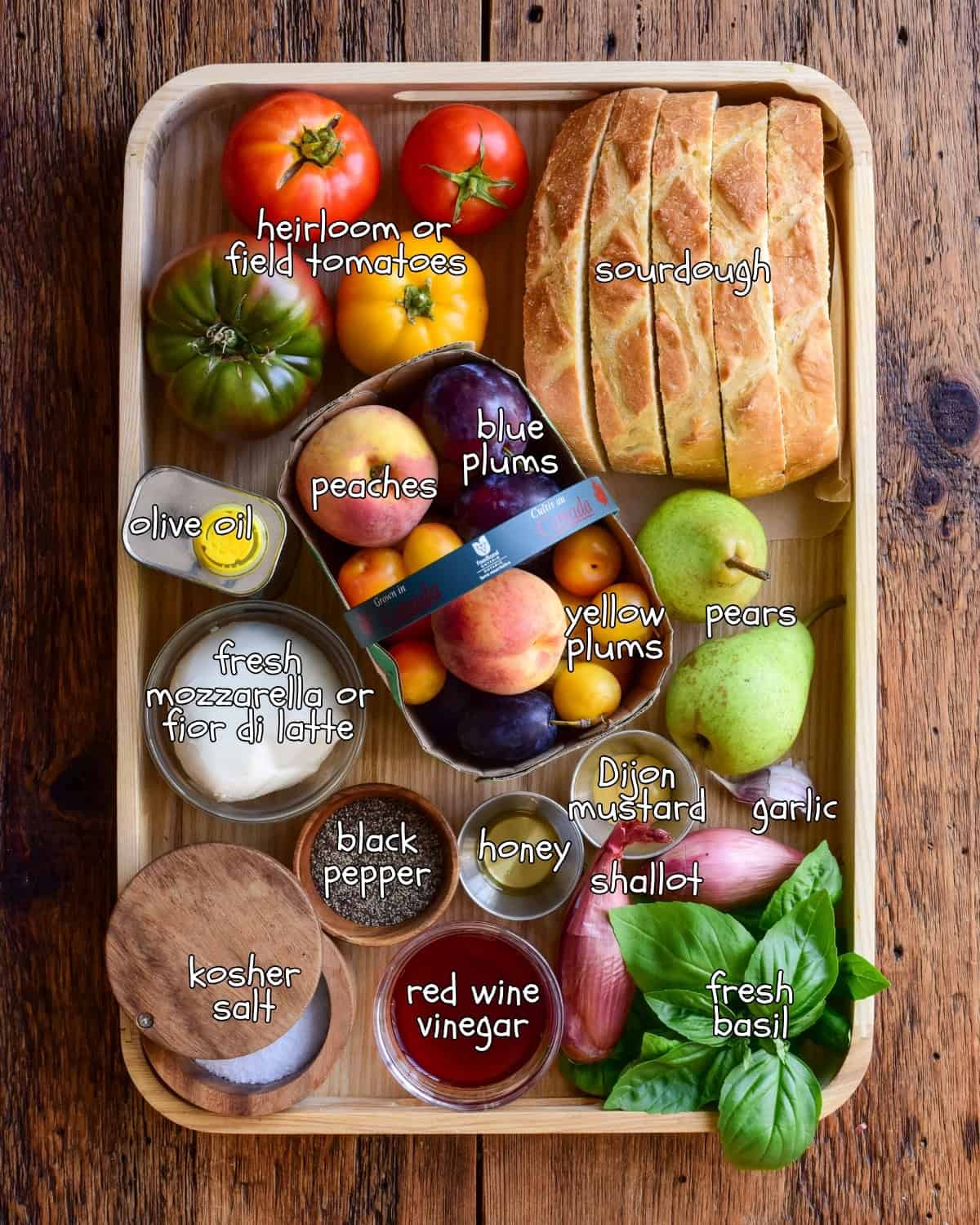 A colourful collection of ingredients for a late-summer stone fruit panzanella recipe in a wooden tray. The ingredients are all labelled.
