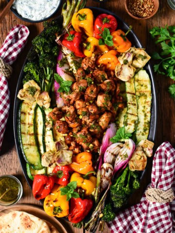 An oval platter with Mediterranean grilled chicken pieces in the center, surrounded by colourful grilled vegetables. Served with a whipped feta tzatziki, grilled flatbreads, drinks and checkered napkins.