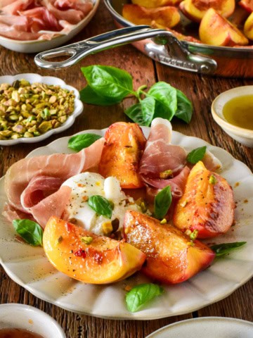 Close-up of pan-seared peaches paired with burrata cheese and prosciutto, garnished with fresh basil leaves, served on a white plate.