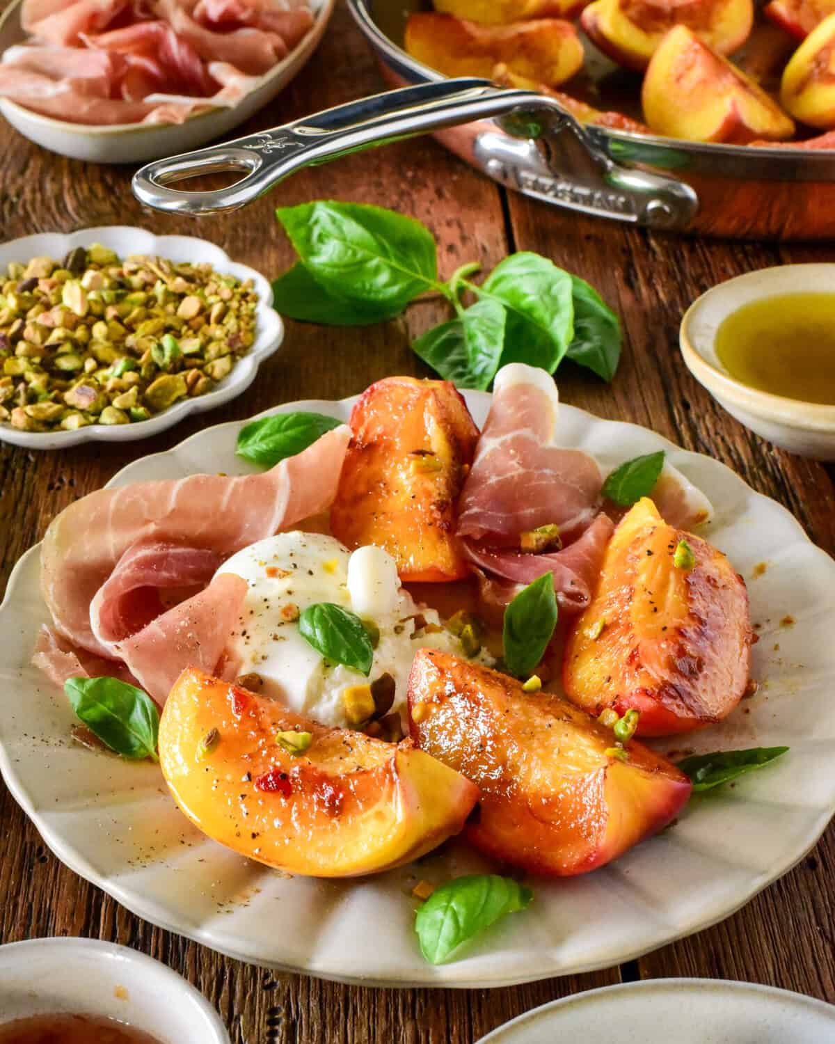 The image features a white plate with slices of pan-seared peaches topped with burrata cheese and prosciutto, garnished with fresh basil leaves. Surrounding the plate are small bowls of pistachios, olive oil, and additional peach slices, set on a rustic wooden table, highlighting a gourmet appetizer inspired by Zimmy's Nook.