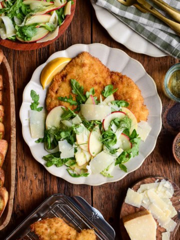 Golden crispy chicken Milanese served with fresh apple fennel salad and lemon wedge on a white plate.