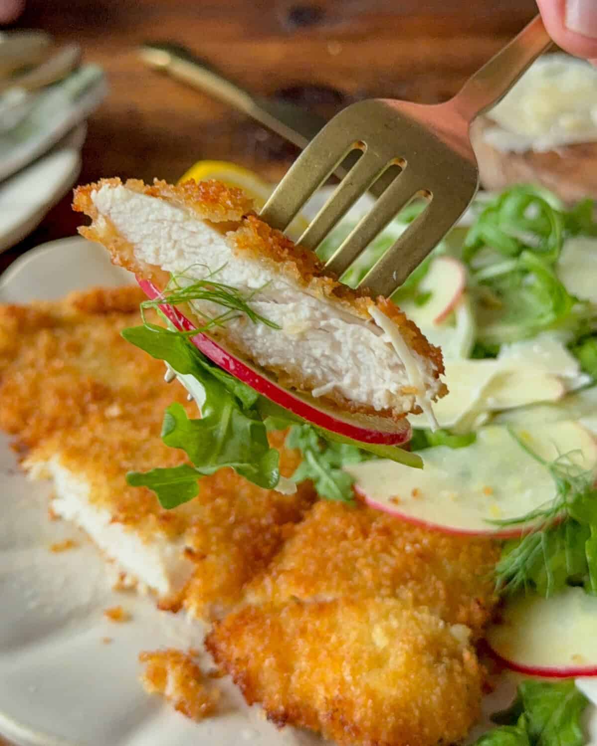 The image features a piece of crispy chicken Milanese on a fork, with a side of fresh apple fennel salad. The chicken has a golden, crunchy breaded coating, and the salad includes sliced apples, fennel, and greens, presented on a white plate.