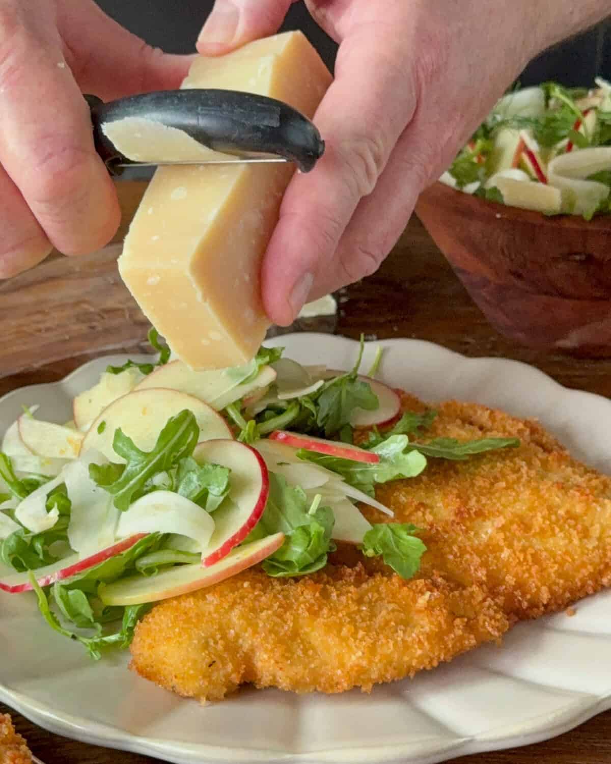 Crispy chicken Milanese served on a white plate with fresh apple-fennel salad and cheese, garnished with microgreens and shaved Parmesan.