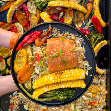 A bowl of miso-maple steelhead trout served over veggie fried rice, surrounded by roasted vegetables including corn, zucchini, and peppers, on a griddle.