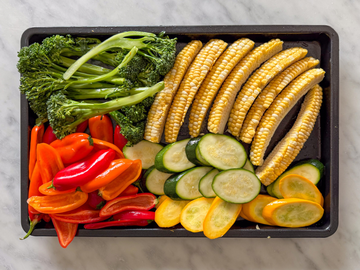 A black-rimmed sheet pan filled with colourful and prepped corn ribs, broccolini, mini peppers and sliced zucchini.