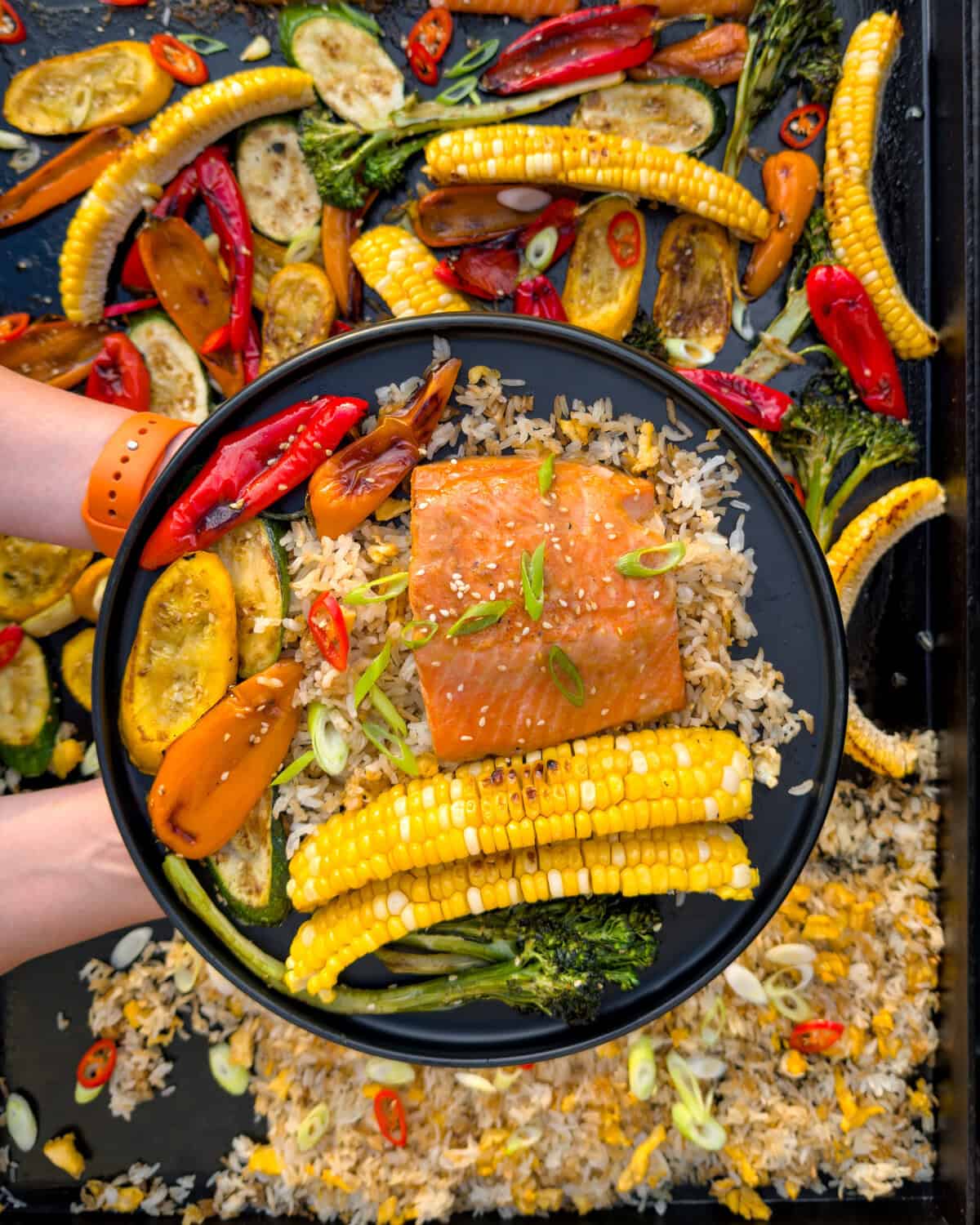 A bowl of miso-maple steelhead trout served over veggie fried rice, surrounded by roasted vegetables including corn, zucchini, and peppers, on a griddle.