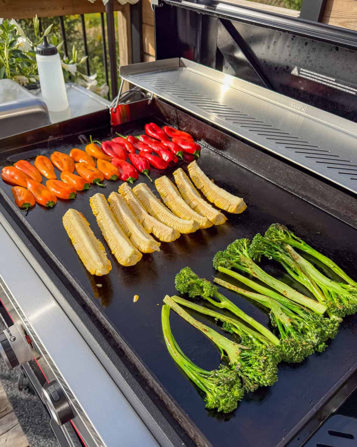 Row of broccolini, corn ribs and mini orange and red peppers on an outdoor griddle.