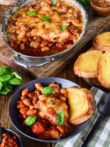 Delicious pizza beans skillet with spicy sausage, melted cheese, and fresh basil, served with toasted garlic bread on a rustic wooden table.