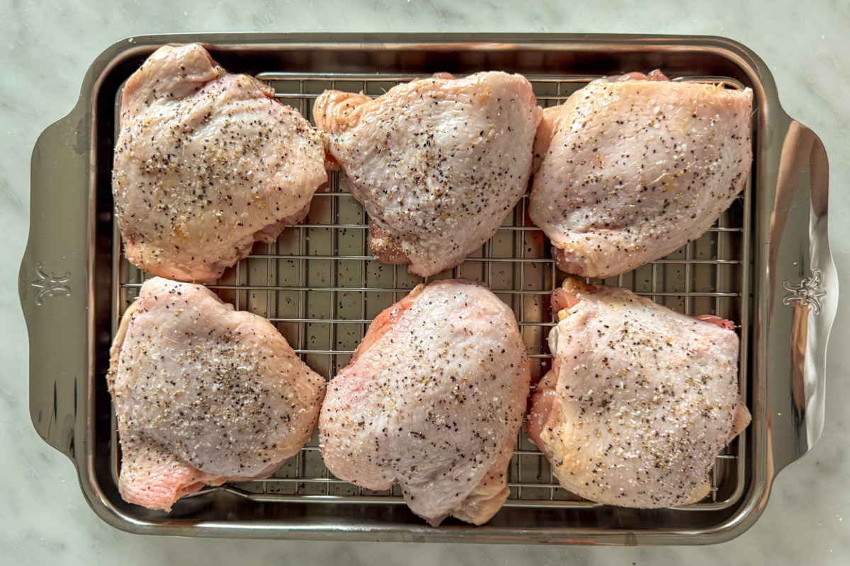 Six skin-on and seasoned chicken thighs on a rack over a sheet pan to dry brine.