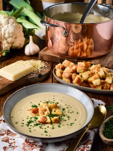 A bowl of creamy roasted cauliflower and leek soup garnished with croutons and chives, served with cheese, garlic, and fresh vegetables on a rustic wooden table.