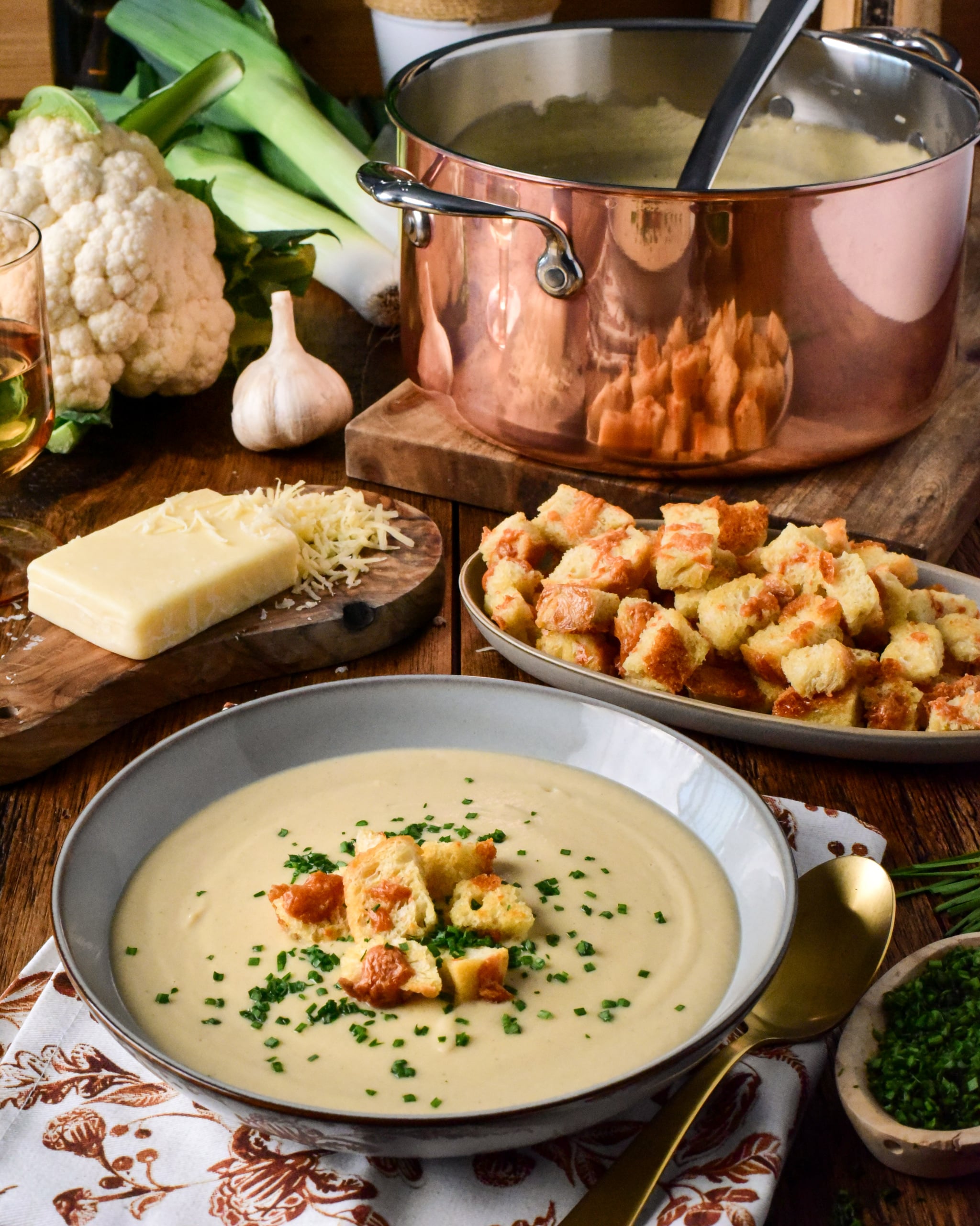 A bowl of creamy roasted cauliflower and leek soup garnished with croutons and chives, served with cheese, garlic, and fresh vegetables on a rustic wooden table.