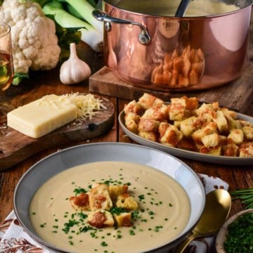 A bowl of creamy roasted cauliflower and leek soup garnished with croutons and chives, served with cheese, garlic, and fresh vegetables on a rustic wooden table.