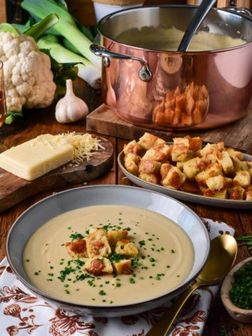 A bowl of creamy roasted cauliflower and leek soup garnished with croutons and chives, served with cheese, garlic, and fresh vegetables on a rustic wooden table.
