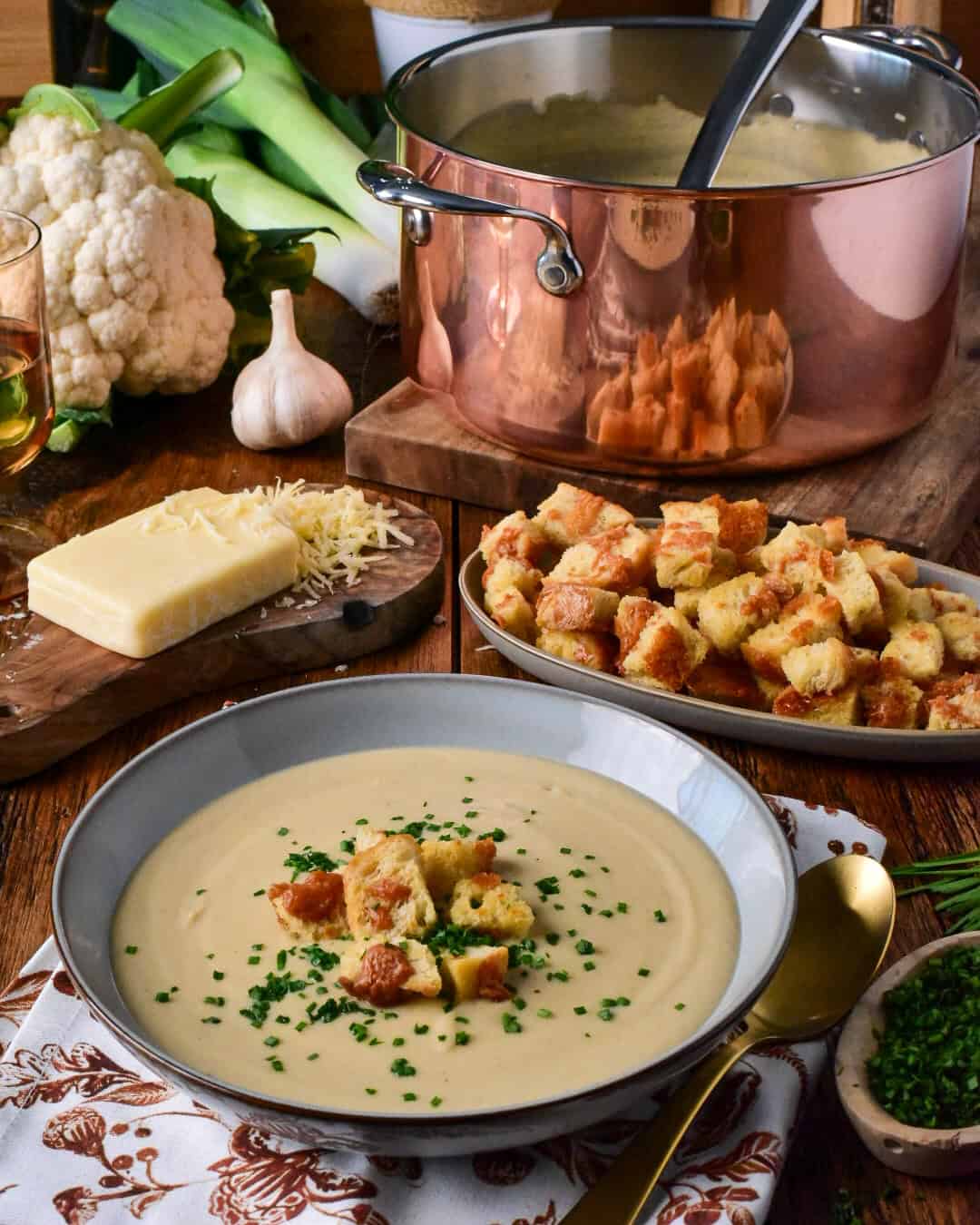 A bowl of creamy roasted cauliflower and leek soup garnished with croutons and chives, served with cheese, garlic, and fresh vegetables on a rustic wooden table.