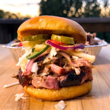 Delicious smoked pulled pork sandwich with pickles, coleslaw, and mustard on a soft bun, served outdoors at sunset.