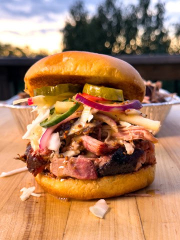 Delicious smoked pulled pork sandwich with pickles, coleslaw, and mustard on a soft bun, served outdoors at sunset.