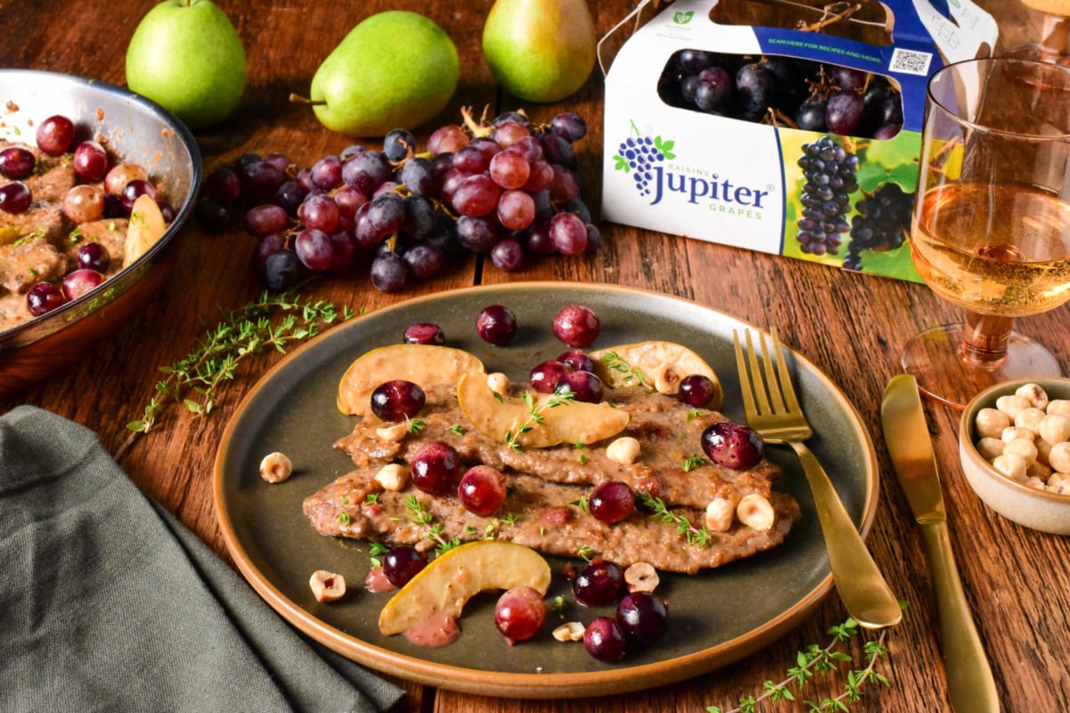 Delicious veal scallopini served with fresh blue grapes and pear slices on a dark plate, garnished with herbs, with a glass of rosé wine in the background.