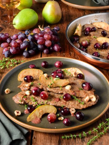 Delicious veal scallopini topped with blue grapes and pear slices, served on a dark plate with fresh grapes and pears in the background.