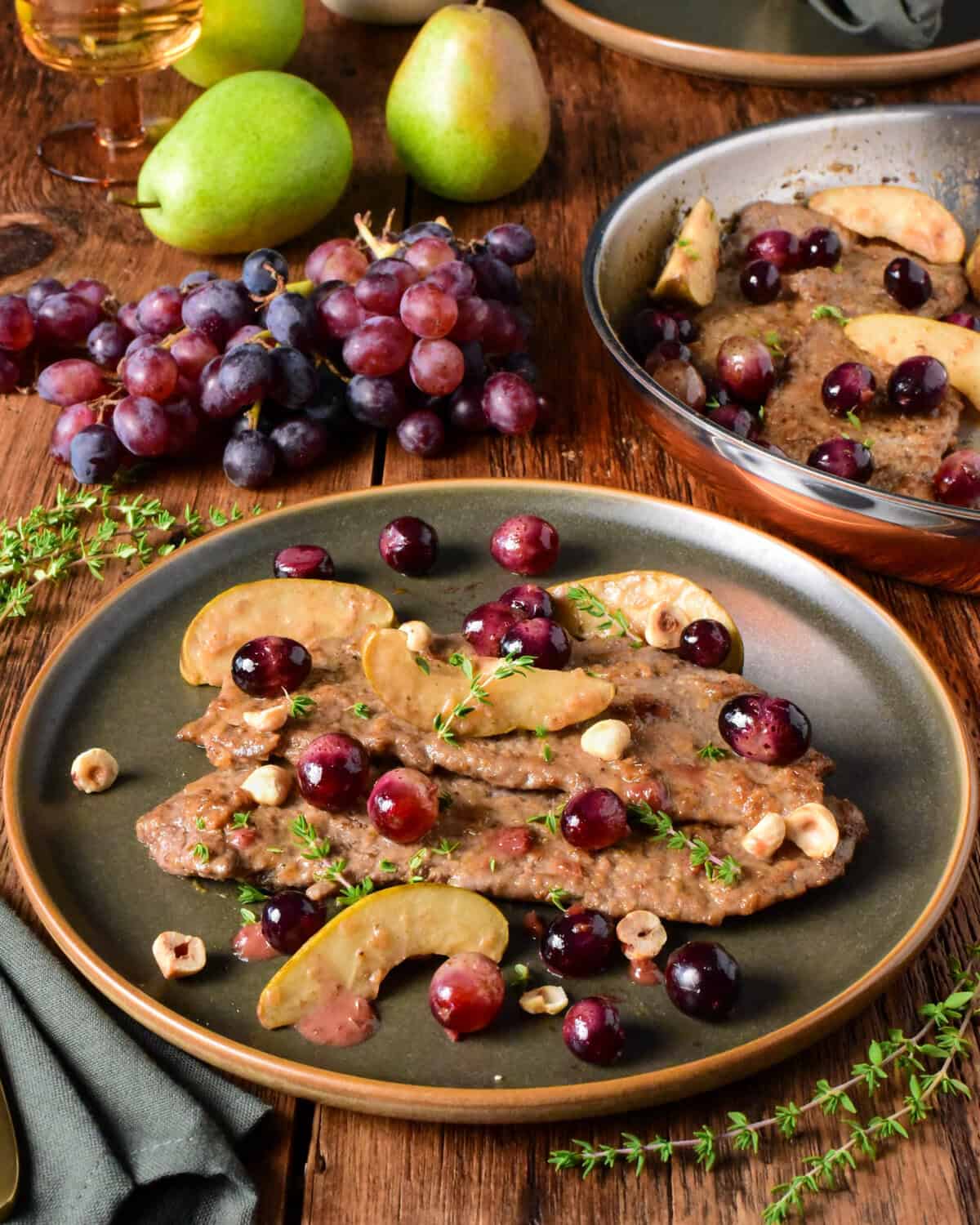 Delicious veal scallopini topped with blue grapes and pear slices, served on a dark plate with fresh grapes and pears in the background.