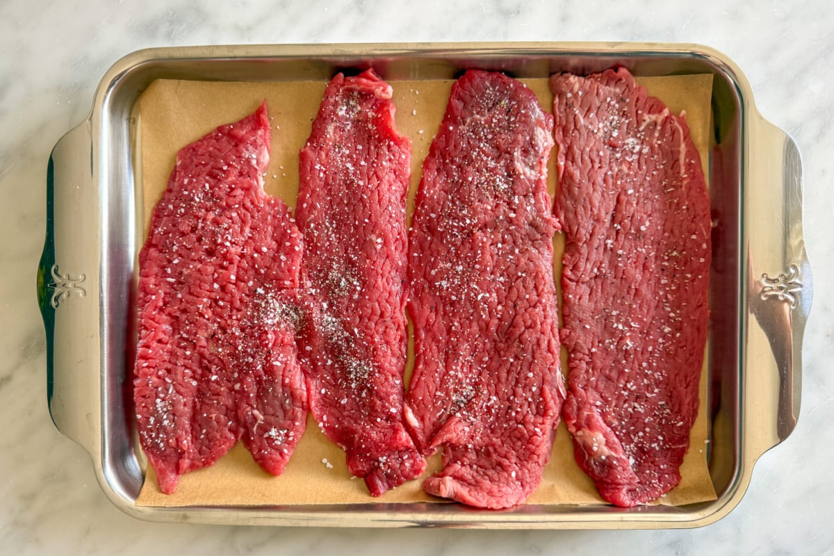 Four pieces of veal on a parchment lined sheet pan, are seasoned with salt and pepper.
