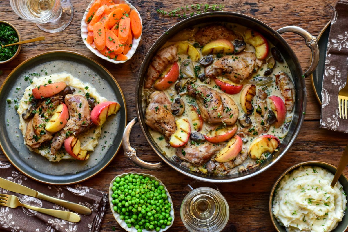 Succulent rabbit braised with apples and vegetables, presented on a rustic plate with herbs, alongside sides of greens and carrot slices.