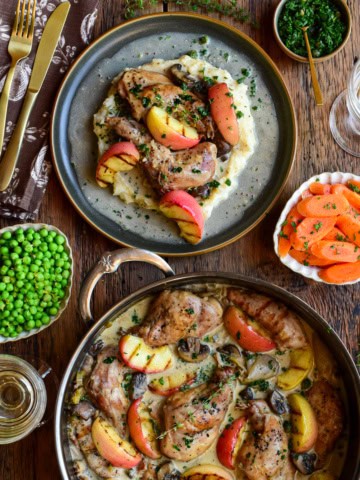 Succulent rabbit braised with apples and vegetables, presented on a rustic plate with herbs, alongside sides of greens and carrot slices.