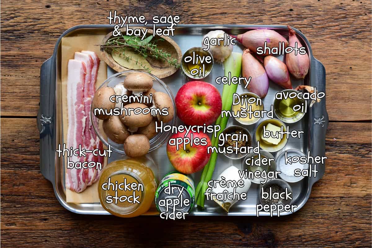 A stainless steel tray with labelled ingredients for rabbit braising: thyme, sage & bay leaf, garlic, shallots, olive oil, celery, Dijon mustard, avocado oil, Honeycrisp apples, apple cider, bacon, chicken stock,.
