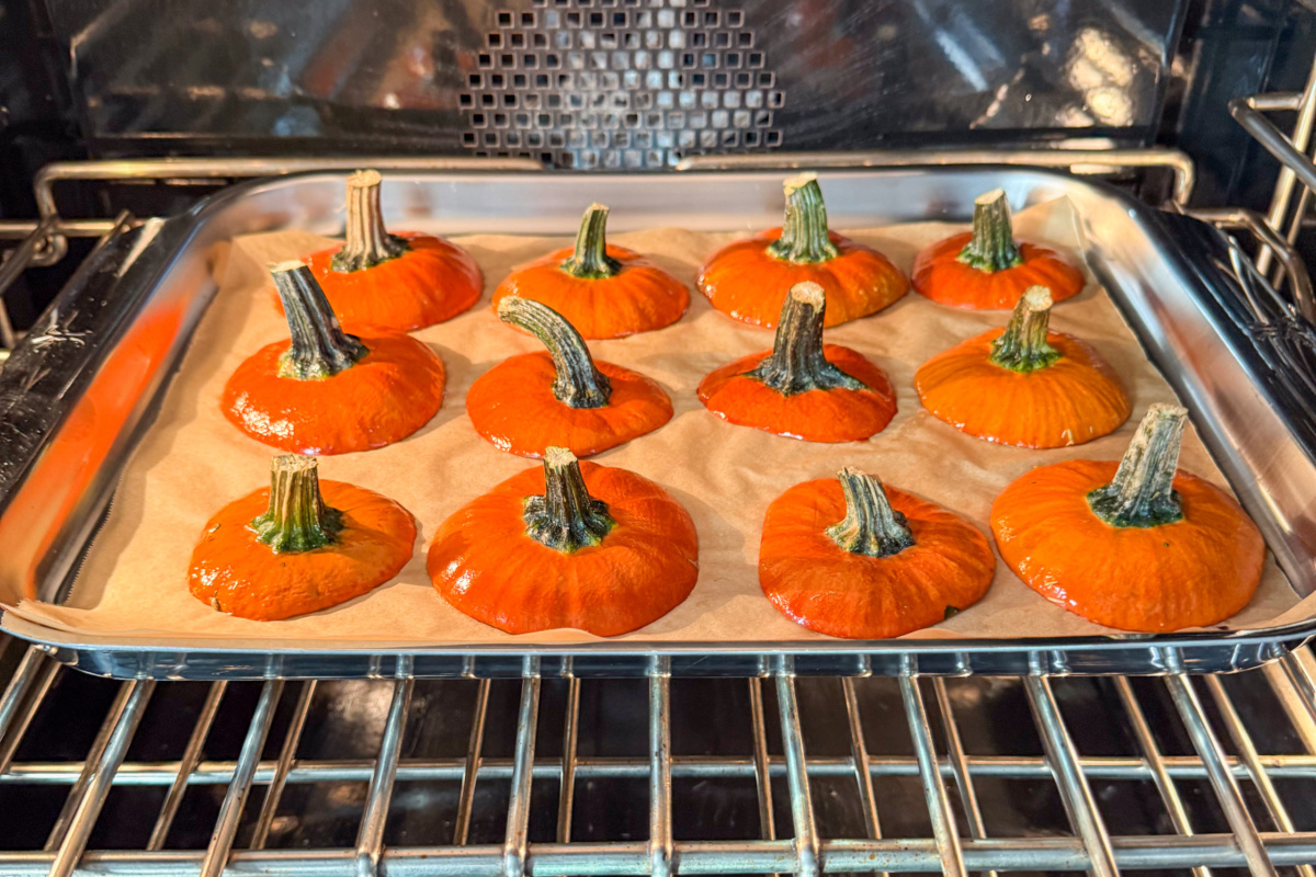 A parchment-lined sheet pan with twelve mini pumpkin lids, in the oven.