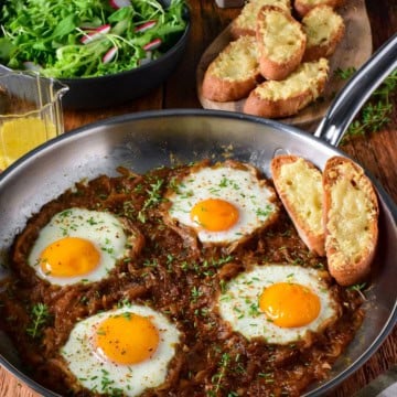 Skillet with baked eggs in caramelized onion and Gruyère cheese, served with toasted bread and fresh salad on a rustic wooden table.