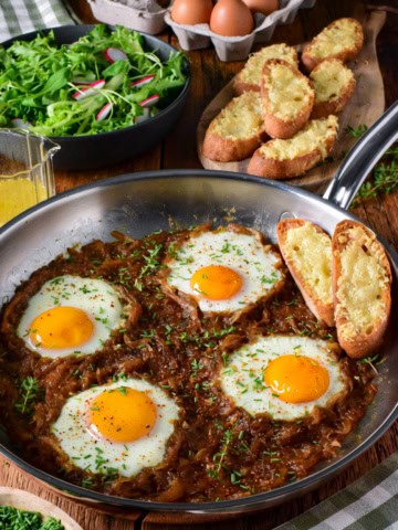 Skillet with baked eggs in caramelized onion and Gruyère cheese, served with toasted bread and fresh salad on a rustic wooden table.